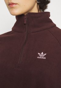 Burgundy fleece-tröja med hög krage, halvdragkedja och vitt Adidas-logotyp. Mjuk textur och subtila sömdetaljer.