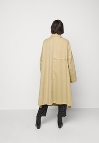 Ljusbeige lång kappa med bakdel och knapprad, gjord av slät tyg. Bärs över svarta rutiga byxor och ankelse352mm.