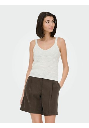 Woman with short dark hair wearing a white knit sleeveless top and dark brown pleated shorts, hands in pockets, looking to the side.