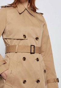 Beżowy płaszcz trencz wykonany z gładkiej tkaniny, z podwójnym zapięciem na guziki, dużymi guzikami, paskiem w talii i bocznymi kieszeniami.