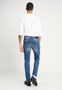 Blue denim jeans with a tapered fit, faded texture, and distinct back pockets. Paired with a white long-sleeve shirt and black shoes.