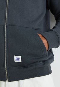 Mørk grå zip-up hettegenser laget av myk stoff, med ribbede mansjetter, frontlommer og en hvit etikett med blå tekst ved kanten.