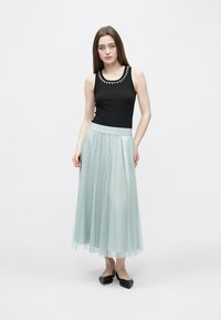 Černý tílko s dekorativním výstřihem, doplněné světlou tyrkysovou, vrstvenou tylovou maxi sukní. Modelka stojí v černých balerínách na jednobarevném pozadí.