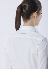 Camicia bianca con bottoni, caratterizzata da una texture liscia, con colletto e dettaglio ricamato sul retro. Capelli raccolti in una coda di cavallo.