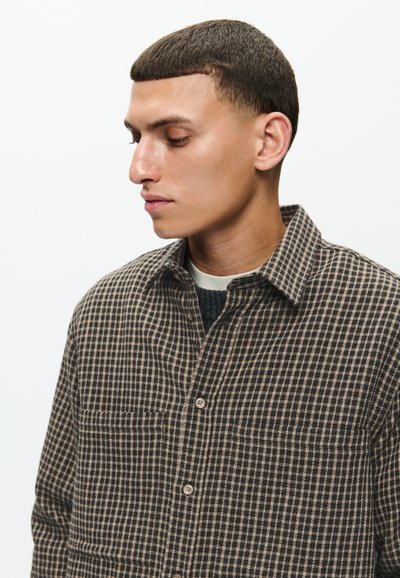 Barna és fekete kockás ing, texturált anyagból készült, gombos nyakkal és elülső zsebekkel, sötét pulóver felett viselve.