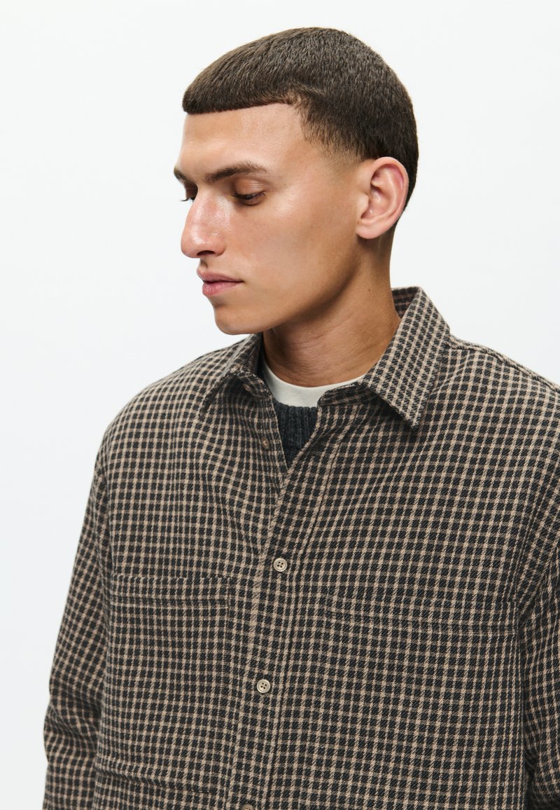 Camicia a quadri marrone e nera realizzata in tessuto texturizzato, con colletto a camicia e tasche frontali, indossata sopra un maglione scuro.