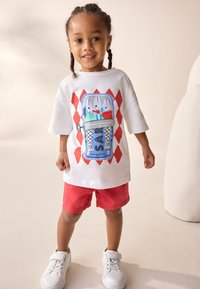 Fehér póló, melyen színes fagylalt grafika látható, piros rövidnadrággal és fehér tornacipővel, egy semleges háttér előtt álló gyermeken.