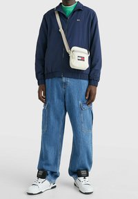 Navy zip-up jacket, oversized blue denim cargo pants, white sneakers with black accents, and a small cream crossbody bag with logo.