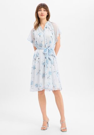 Kobieta w białej sukience do kolan z niebieskim kwiatowym wzorem, krótkimi falującymi rękawami, przewiązaną talią i srebrnymi sandałami na wysokim obcasie stojąca na białym tle.