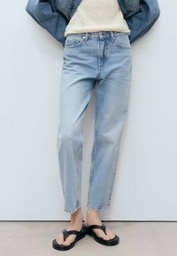Jeans de mezclilla de un azul claro con un ajuste relajado, piernas rectas y cintura alta. Llevados con sandalias de dedo negras sobre un fondo neutro.