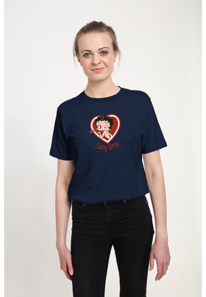Femme portant un t-shirt bleu marine avec un motif de Betty Boop à l'intérieur d'un cœur rouge et un pantalon noir, debout sur un fond blanc.