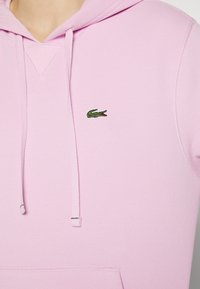 Sudadera con capucha de color rosa claro, confeccionada en tejido texturizado, con capucha ajustable mediante cordón y un pequeño logo de cocodrilo verde en el pecho.