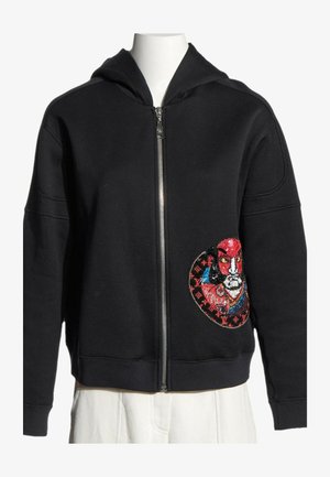 Sweat à capuche zippé noir avec un patch visage brodé coloré sur la partie inférieure droite, présenté sur un torse de mannequin blanc.