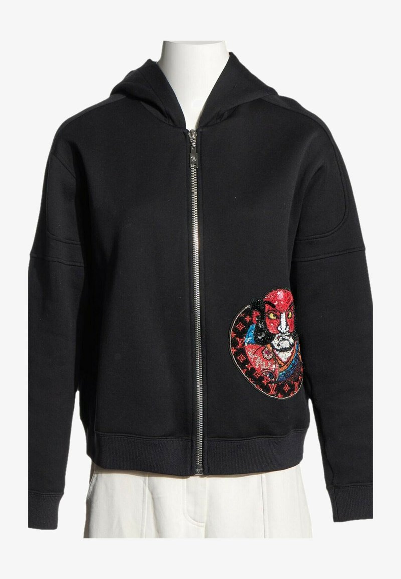 Sweat à capuche zippé noir avec un patch visage brodé coloré sur la partie inférieure droite, présenté sur un torse de mannequin blanc.