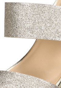 Correa superior plateada brillante con un acabado texturizado, combinada con una plantilla beige suave, que presenta una punta abierta y un diseño minimalista.