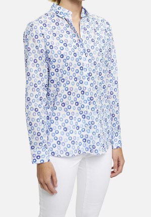 Chemise blanche boutonnée avec un motif circulaire bleu et violet clair. Manches longues, col et ourlet droit. Portée avec un pantalon blanc.