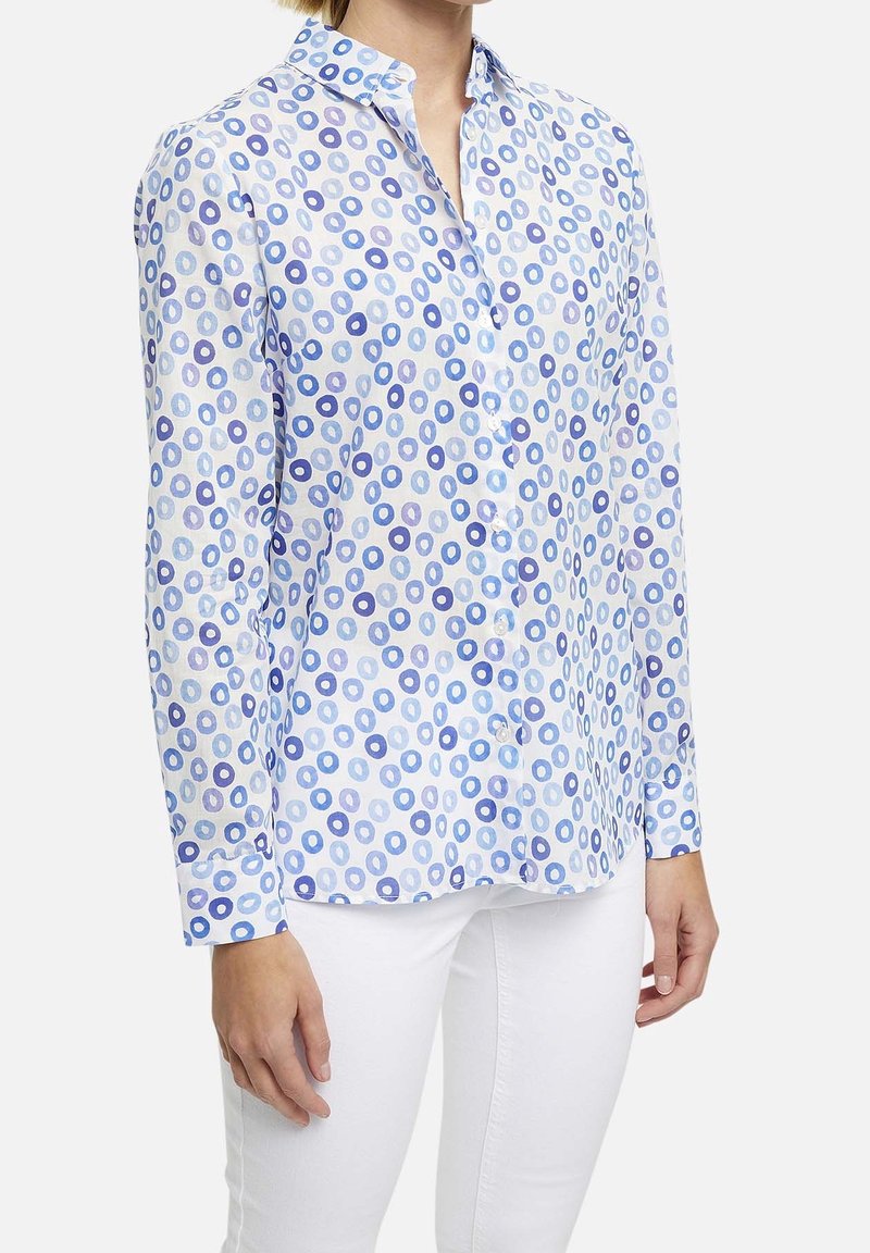 Chemise blanche boutonnée avec un motif circulaire bleu et violet clair. Manches longues, col et ourlet droit. Portée avec un pantalon blanc.