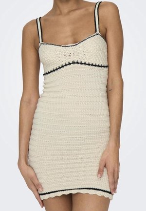 Femme portant une robe au crochet sans manches de couleur crème avec un bord noir le long de l'encolure, des bretelles et de l'ourlet.