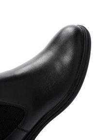 Zapato de cuero negro con punta redonda, panel lateral de malla elástica y superficie texturizada. Presenta una suela de goma negra resistente.