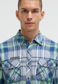 Karierter blau-grüner Flanellhemd mit zwei Brusttaschen, Knopfverschluss und einem Markenetikett an der linken Tasche.