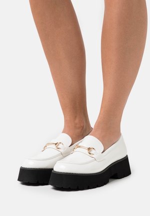 White patent leather loafers with a chunky black platform sole, featuring a gold chain accent across the vamp and a smooth texture.