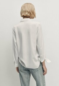 Vaaleahiuksinen henkilö, jolla on pitkähihaista valkoista paitaa, jossa on sidottu hihansuut, sekä vaaleansiniset farkut, kääntynyt pois yksinkertaista vaaleata taustaa vasten.