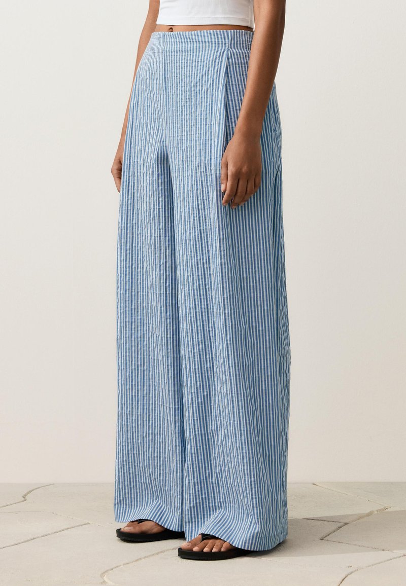 Femme portant un pantalon taille haute à jambes larges rayé verticalement bleu et blanc, avec des sandales noires et un top court blanc.