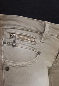 Los jeans de mezclilla beige cuentan con un bolsillo con cremallera para monedas, acentos de ferretería metálica y detalles de costura sutiles. Textura suave y diseño ajustado.