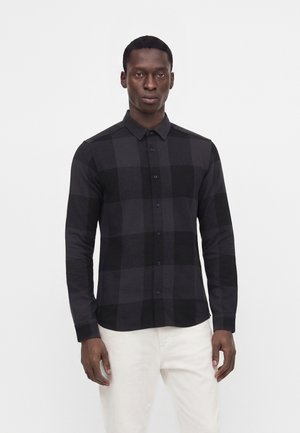 Homme portant une chemise à carreaux noire et gris foncé à boutons avec manches longues et pantalon blanc, debout devant un fond clair uni.