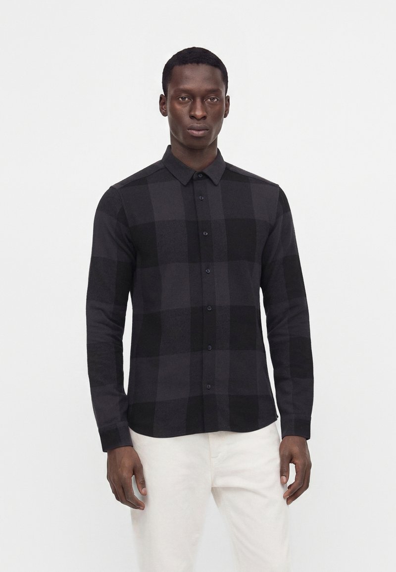 Homme portant une chemise à carreaux noire et gris foncé à boutons avec manches longues et pantalon blanc, debout devant un fond clair uni.