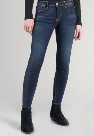 Dunkelblaue, enge Jeans aus Denim mit mittelhohem Bund, klassischem Fünf-Taschen-Design und kontrastierenden Nähten. Getragen mit schwarzen Stiefeletten mit Absatz.