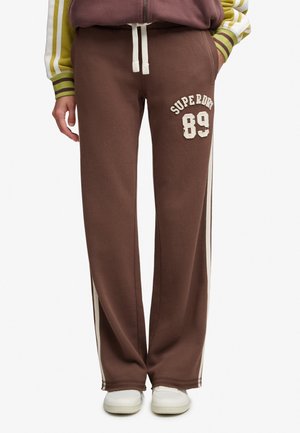 Brune joggingbukser med hvide sidelister, hvid snøre og "Superdry 89" tekst på den øverste venstre ben, båret med hvide sneakers.