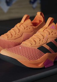 Paire de chaussures de sport orange et rose avec des bandes noires, texture en mesh et semelles épaisses et rembourrées, posées sur une surface sombre.