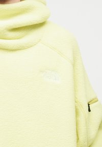 Ανοιχτό κίτρινο λεπτό φλις με ψηλό γιακά και μικρή τσέπη με φερμουάρ στο αριστερό μανίκι, με κεντημένο λευκό λογότυπο "The North Face" στο στήθος.