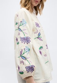 Femme portant un chemisier crème à manches longues bouffantes brodées de motifs floraux violets, bleus et verts.