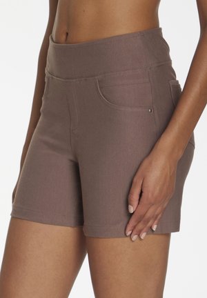 Close-up of a person wearing high-waisted, brown fitted shorts with front and side pockets, standing against a plain background.