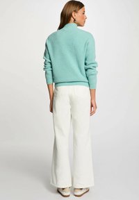 Maglione lavorato a maglia turchese chiaro con colletto alto, abbinato a jeans bianchi a gamba larga e sneakers chiari, visto da dietro.