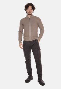 Maglione beige con zip e texture a coste, abbinato a pantaloni cargo scuri con molteplici tasche. Stivaletti neri completano l'outfit.