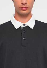 Svart polo-skjorte med hvit krage, med tre knapper. Stoffet er glatt med en liten tekstur. Klassisk avslappet design.