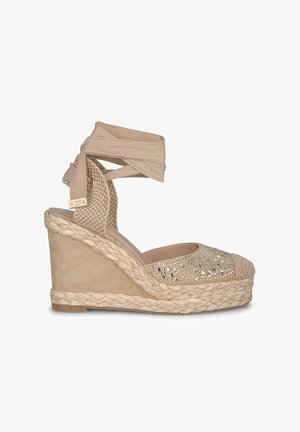 Nude-Wedge-Espadrille mit geflochtener Jutesohle, Obermaterial aus strukturiertem Stoff, verziert mit metallischen Nieten und Riemen für einen Wickelstil.