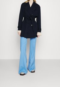 Tmavě modrý trenčkot s páskem a dvouřadými knoflíky, vrstvený přes světle blůzu a doplněný světle modrými rozšířenými kalhotami a kovovými sandály.