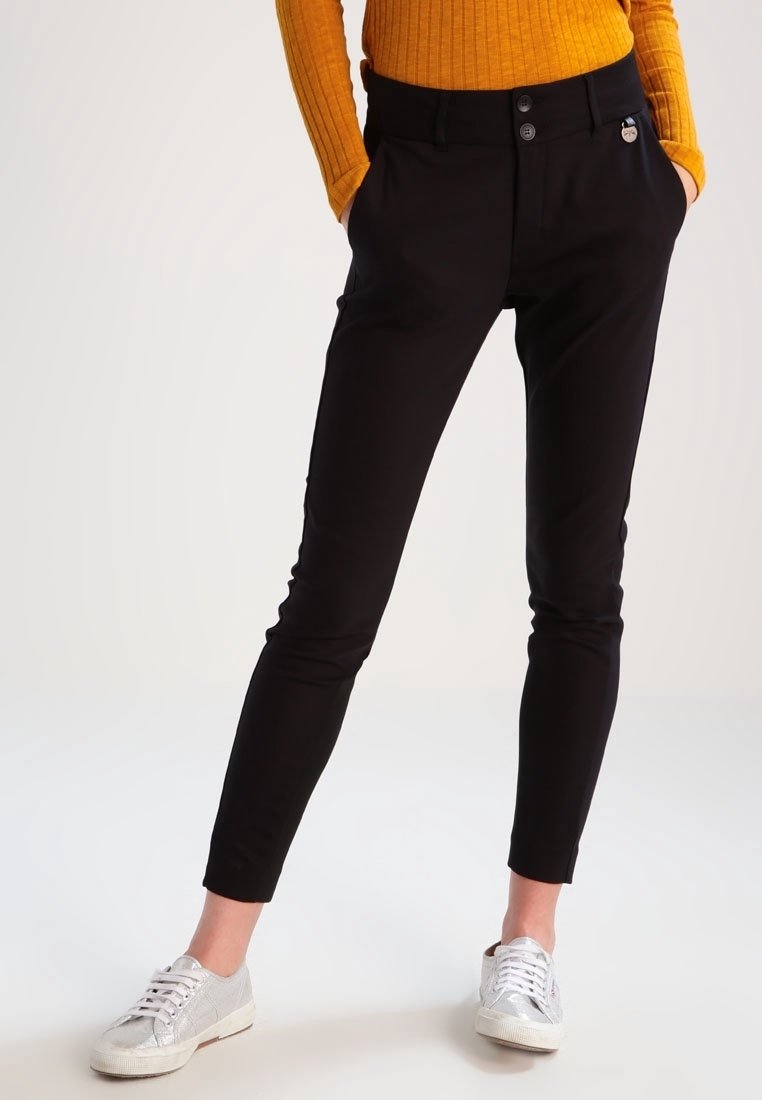 Personne portant un pantalon slim noir, un haut à manches longues côtelé orange et des baskets argentées à lacets, debout les mains dans les poches.
