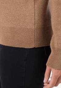 Maglione a maglia color tan con orlo e polsini a coste, mostrato insieme a jeans neri. Il tessuto appare morbido con una texture liscia.