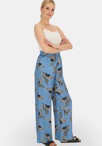 Niebieskie spodnie o szerokich nogawkach z czarno-białym wzorem liści, z elastycznym paskiem w talii i gładką teksturą materiału. Połączone z białą bluzką.