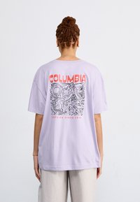 Maglietta oversize lavanda con un design grafico di montagne e la scritta "COLUMBIA" in rosso sul retro. Materiale in cotone.