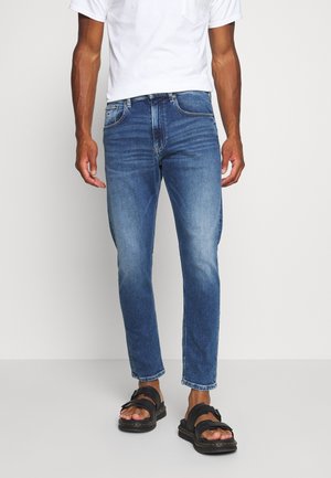 Man wearing mid-wash slim-fit jeans, white short-sleeve shirt, and black open-toe sandals standing against a plain background.