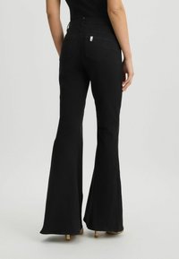 Jeans neri svasati con vita alta, caratterizzati da una tasca posteriore e tessuto strutturato, mostrati da dietro, abbinati a tacchi eleganti.