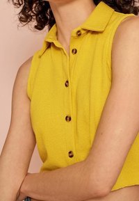 Modelo vistiendo una blusa sin mangas de color amarillo brillante con cuello y detalles de botones, de pie frente a un fondo rosa suave.