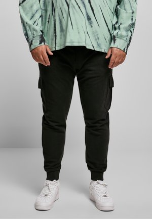 Person trägt schwarze Cargohose, weiße Sneakers und ein grünes Batik-Langarmshirt, steht vor grauem Hintergrund.