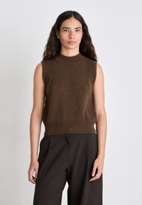 Brauner ärmelloser Strickpullover mit hohem Kragen und geripptem Saum. Getragen mit dunklen, weit geschnittenen Hosen. Einfaches und sauberes Design.